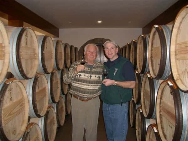Two men toasting with red wine. In the background are wood barrels.