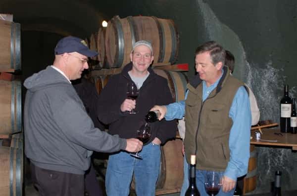 Three men taste testing red wine. In the background are wood barrels.