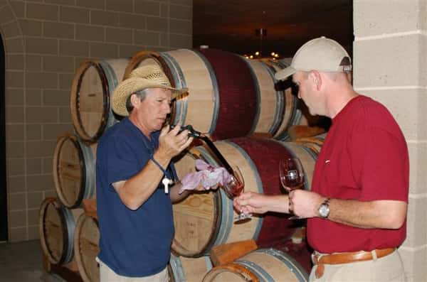 Two men taste testing red wine. In the background are wood barrels.