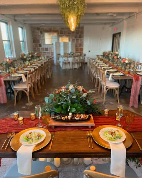 A beautifully decorated event space featuring long tables set for a wedding or corporate catering, adorned with floral centerpieces and elegant tableware, creating a warm ambiance.