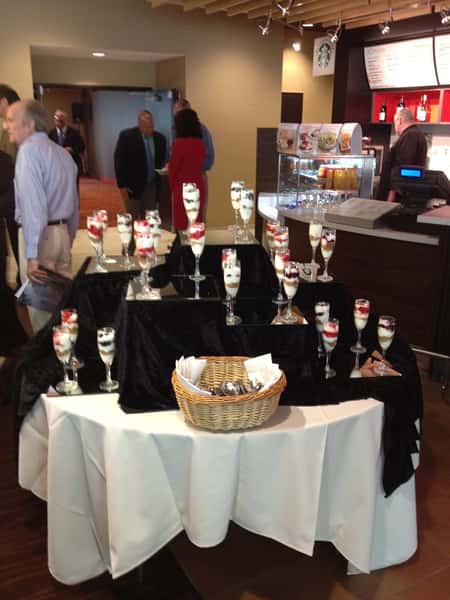 A catering display features elegant parfaits in tall glasses, layered with yogurt, fruit, and granola, set on a black tablecloth.
