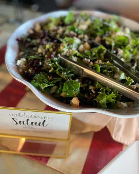 A fresh autumn salad featuring mixed greens, nuts, and feta cheese, served in a white bowl with tongs. A sign indicates "Autumn Salad with Balsamic Dressing," perfect for catering.