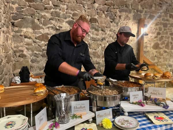 Two chefs in black uniforms serve food at a rustic catering event, featuring pulled pork, chicken, and sliders.