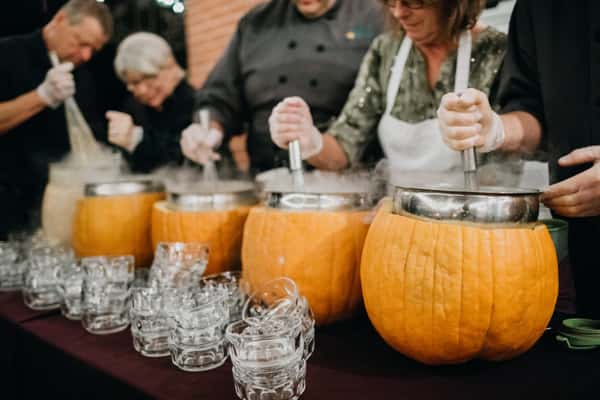 Caterers prepare a unique pumpkin soup in hollowed-out pumpkins, with steam rising, while guests enjoy the festive atmosphere, showcasing creativity in event catering.