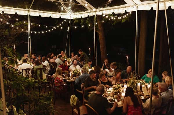 A beautifully decorated outdoor dining setup under a tent, featuring guests enjoying a meal with soft lighting, floral centerpieces, and a warm, inviting atmosphere for a special event.