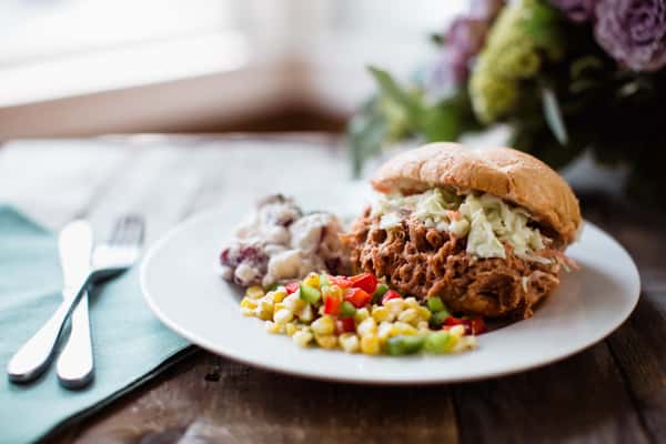 A delicious pulled pork sandwich topped with coleslaw, served alongside creamy potato salad and a colorful corn salad, perfect for barbecue catering at weddings or corporate events.