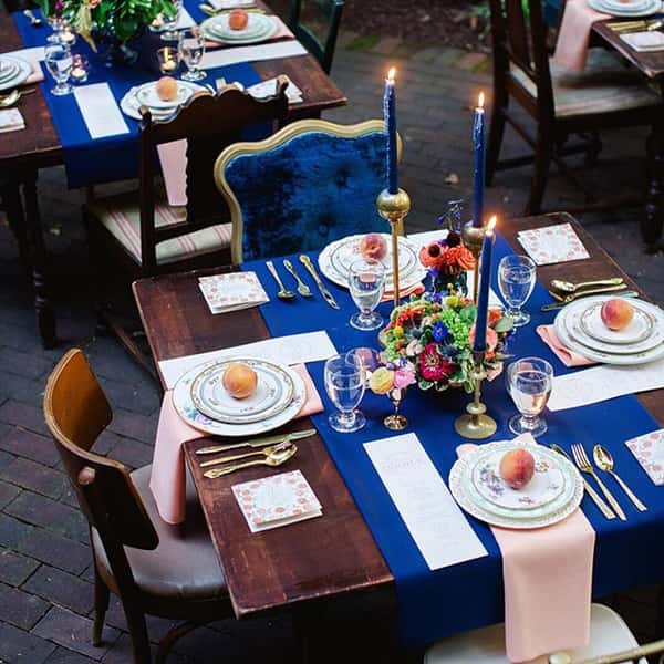 An elegant catering setup featuring a beautifully arranged table with navy and blush linens, floral centerpieces, fine china, and candles, perfect for a sophisticated outdoor event.