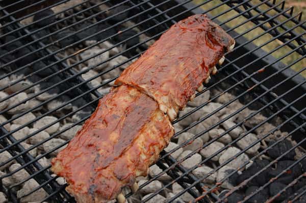 A rack of barbecue ribs glistens with sauce as it cooks on a grill over hot coals.