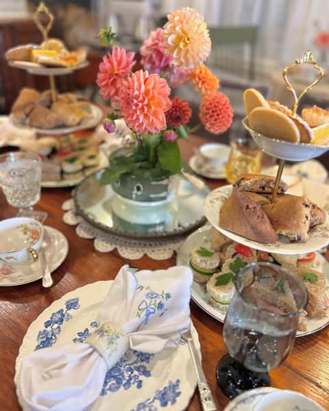 A charming tea party setting featuring a tiered stand with assorted pastries and sandwiches, vibrant dahlias in a vase, elegant china, and a beautifully folded napkin, perfect for gatherings.