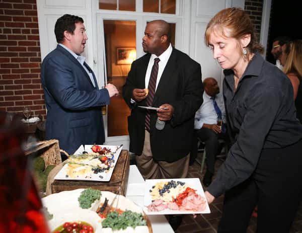 Two professionals engage in conversation at a corporate catering event, while a server presents a platter of meats and cheeses, enhancing the elegant atmosphere with delicious offerings.