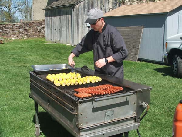 A chef in a black uniform grills hot dogs and corn on a large outdoor grill, set against a green lawn and rustic buildings, ideal for catering events and barbecues.