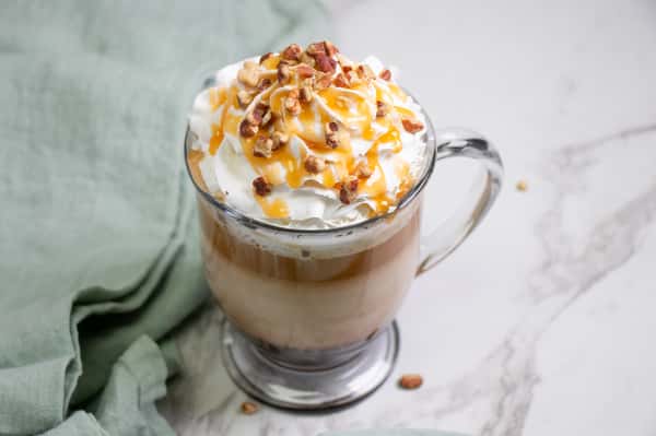 A caramel coffee drink topped with whipped cream, drizzled with caramel sauce, and sprinkled with crushed nuts in a glass mug.