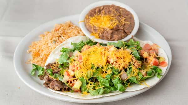 A plate with two soft tacos filled with seasoned ground beef, lettuce, tomatoes, and shredded cheddar cheese, served with Mexican rice and refried beans.