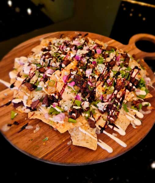 Loaded nachos topped with diced steak, red onions, green onions, crumbled cheese, and drizzled sauces on a wooden platter.