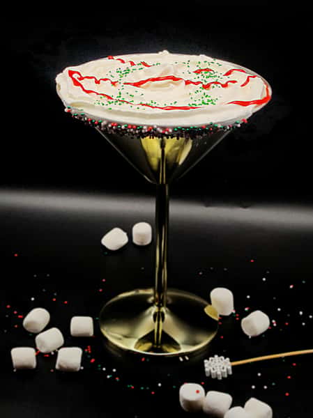 Festive martini topped with whipped cream, red drizzle, green sprinkles, and rimmed with colorful candy beads, surrounded by marshmallows.