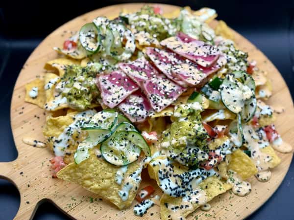 Fusion nachos topped with cucumber slices, salami, guacamole, creamy drizzle, and black sesame seeds on a wooden board.