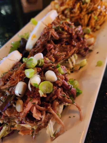 Bao buns filled with shredded meat, slaw, and topped with chopped green onions on a rectangular white plate.