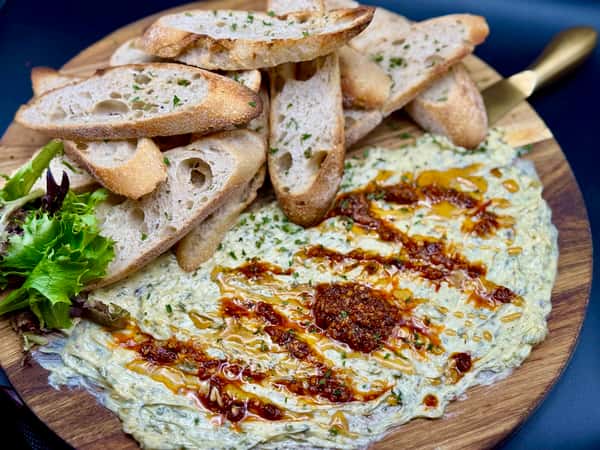 Creamy spinach dip topped with chili oil, served with toasted baguette slices on a wooden board with fresh greens.