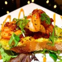 Grilled shrimp atop toasted bread with greens and drizzle of sauce, served on a square white plate with a decorative garnish.