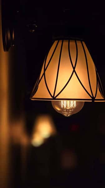 Warmly lit vintage wall sconce with a decorative glass shade and exposed bulb, glowing against a dark background.
