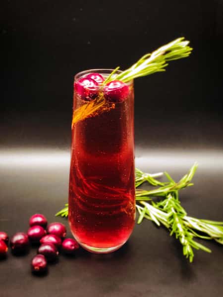 This festive drink appears to be a sparkling cranberry cocktail or mocktail, garnished with fresh cranberries and a sprig of rosemary, giving it a holiday-themed presentation. The glass's tall flute style suggests it's likely a fizzy drink, possibly using champagne, prosecco, or a sparkling water base.
