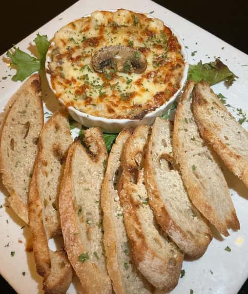 Baked cheesy mushroom dip topped with herbs, served with toasted baguette slices on a square white plate.