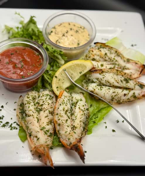 Grilled split lobster tails garnished with herbs, served with cocktail and aioli sauces on a bed of lettuce and a lemon wedge.