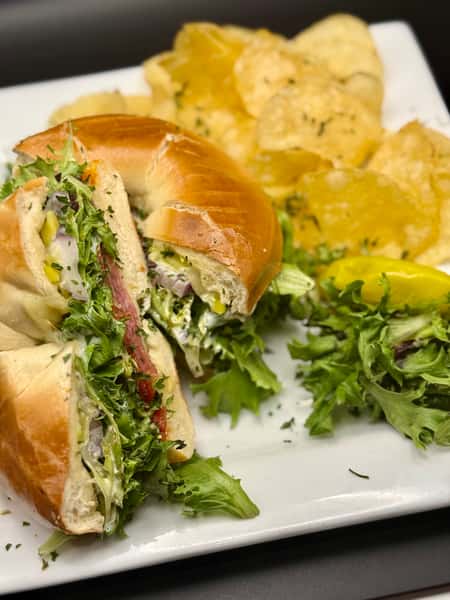 Bagel sandwich filled with fresh greens, onions, and deli meat, served with potato chips and a pepperoncini on a white plate.
