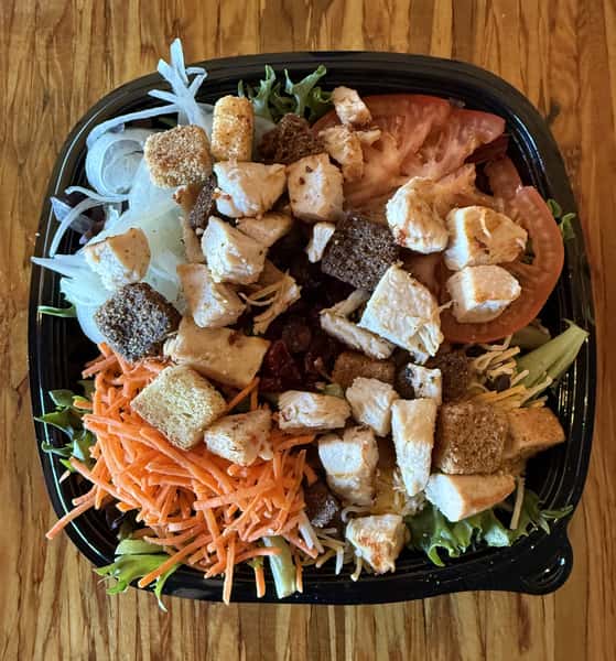 A salad with chicken, croutons, tomatoes, shredded carrots, and lettuce is presented in a black bowl from Main Street Eats.