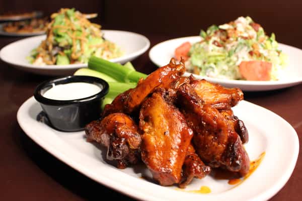 Plate of glazed chicken wings with celery and dipping sauce, alongside salads in the background