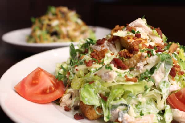 Salad topped with fried chicken and bacon, drizzled with dressing and served with a tomato wedge
