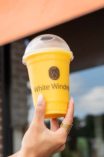 A hand holding a bright yellow fruit smoothie in a clear branded "White Windmill" cup, captured outdoors with a modern café backdrop.
