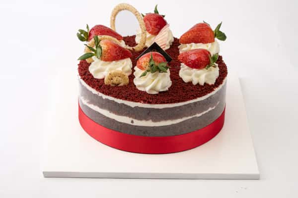 Strawberry red velvet layer cake topped with fresh strawberries and whipped cream dollops, finished with a red ribbon base for an elegant bakery dessert presentation.