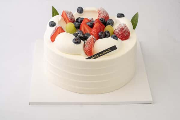 Fresh mixed berry cream cake topped with strawberries, blueberries, grapes, and whipped cream dollops, creating a light and elegant bakery dessert presentation.
