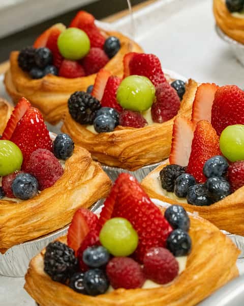 A close-up of individual fruit danishes made with flaky puff pastry, each filled with cream and topped generously with fresh fruit. The toppings include strawberries, blueberries, raspberries, blackberries, and green grapes, arranged in small tart-like pastry shells.