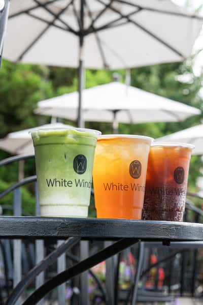 Three iced beverages—green matcha latte, orange fruit drink, and dark iced tea—served in clear cups on an outdoor café table with patio umbrellas in the background.