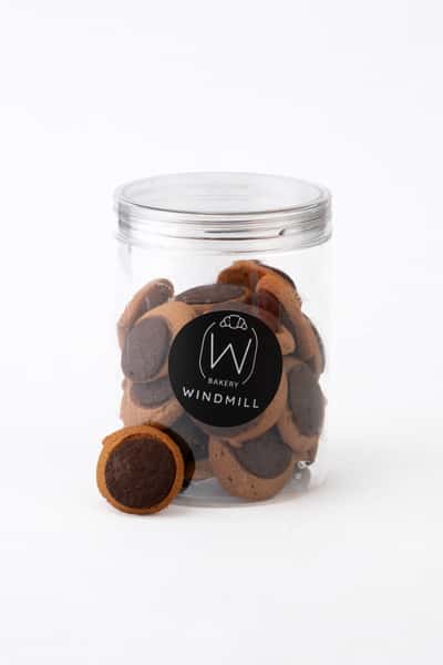 Chocolate sandwich cookies in a clear branded bakery jar, featuring bite-sized cocoa cookies with dark chocolate centers, styled as a gourmet packaged dessert on a clean white background.