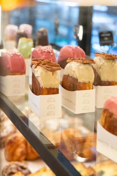 Assorted gourmet croissant pastries with icing and toppings displayed in a bakery case, featuring chocolate drizzle, glaze, and colorful artisan finishes in a modern café setting.