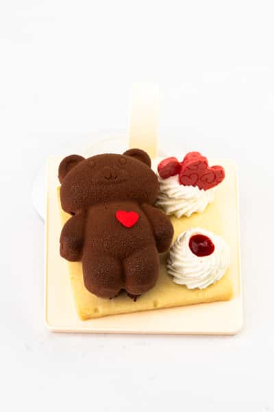 Chocolate bear-shaped mousse cake with velvety finish, decorated with a red heart accent, whipped cream, and berry garnish, styled as a cute gourmet bakery dessert on a clean background.