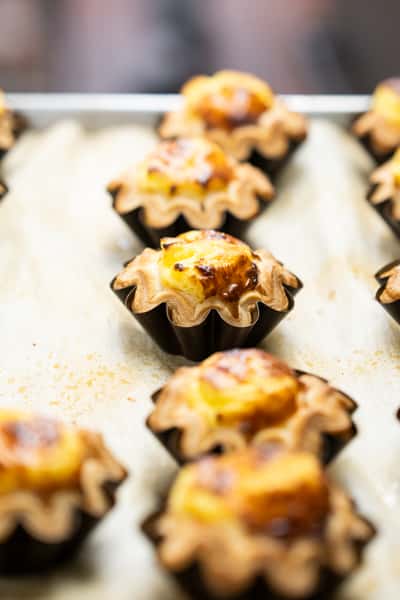 Mini baked egg custard tartlets with caramelized tops in fluted pastry shells, arranged on a baking tray for a fresh bakery display.