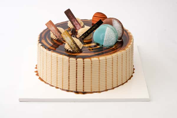 Caramel glaze cake with glossy swirl topping, decorated with macarons, chocolate shards, and cream dollops for an elegant bakery dessert display.