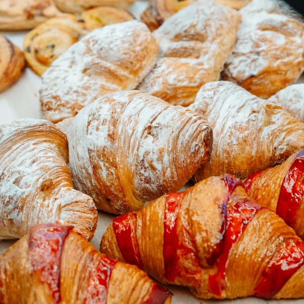 A close-up of assorted croissants and pastries on display. Some are dusted generously with powdered sugar, while others are topped or filled with bright red fruit glaze, likely strawberry.