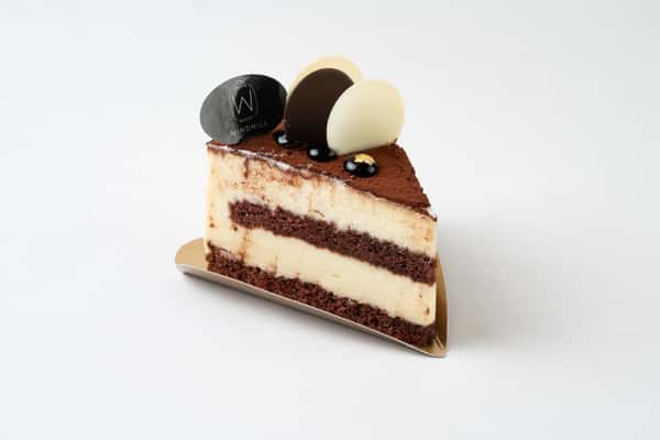 Slice of layered chocolate and vanilla mousse cake with cocoa-dusted top, decorated with chocolate discs and elegant bakery-style presentation on a minimalist background.