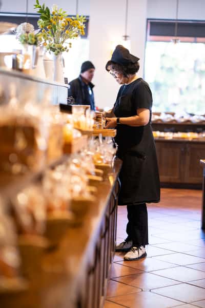 Bakery staff member preparing fresh goods behind the counter in a bright café, with artisanal packaged items and warm, inviting interior ambiance.