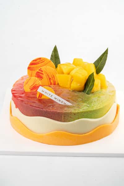 Colorful mango and matcha mousse cake with glossy mirror glaze, topped with fresh mango cubes, decorative chocolate discs, and edible leaves, presented as a modern gourmet bakery dessert.