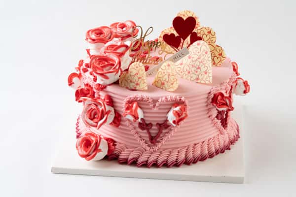 Valentine's Day themed pink buttercream cake decorated with piped roses, heart accents, and white chocolate toppers, creating a romantic artisan bakery dessert centerpiece.