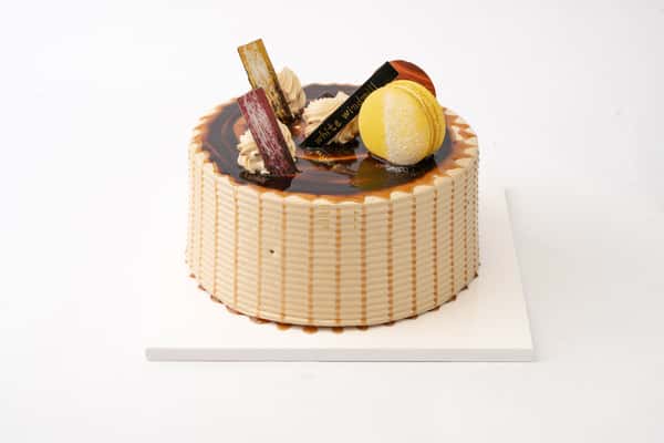 Caramel glaze cake with glossy swirl topping, decorated with macarons, chocolate shards, and cream dollops for an elegant bakery dessert display.