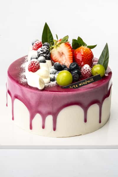 Berry drip cake with glossy purple glaze, topped with fresh strawberries, blueberries, raspberries, green grapes, and whipped cream, styled as an elegant artisan bakery dessert.