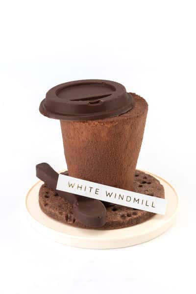 Chocolate coffee cup–shaped dessert with velvety cocoa coating and chocolate lid detail, served with a biscuit base and chocolate garnish, styled as a creative artisan café dessert.