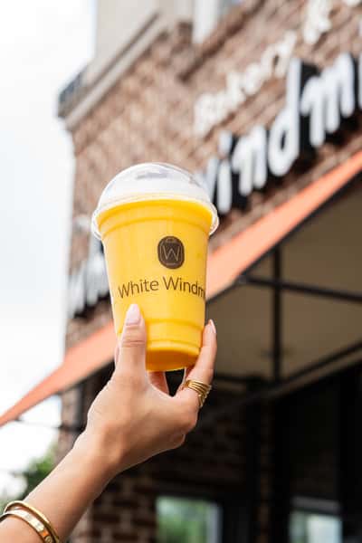 Hand holding a yellow smoothie in a clear plastic cup from "White Windmill Bakery" with a clear dome lid.